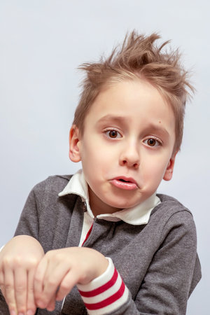 Young boy with tousled hair making a playful face, wearing a gray collared shirt with red and white...