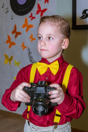 Young child wears a red shirt and yellow bow tie, holding a camera. Wall behind has colorful butterflies. Light decoration scene. Focused expression, bright indoor lightingの写真素材