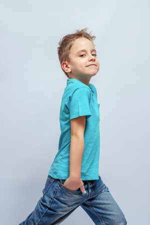 Young boy in a blue shirt and jeans walks confidently with one hand in his pocket, smiling slightly. His hair is styled casually, and he exudes a playful, carefree attitudeの写真素材