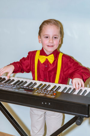 Young child in red shirt with yellow bow tie and suspenders plays an electronic keyboard on a black stand, smiling confidently. Light background highlights cheerful atmosphereの写真素材