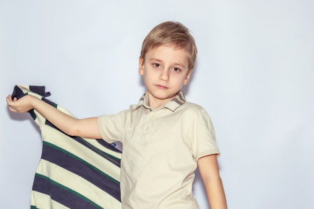 Young boy with short blond hair in a light polo shirt holds a striped shirt in his left hand, gazing confidently at the camera. His expression is calm and composedの写真素材