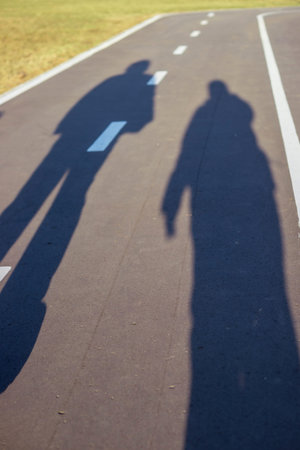 Shadow of two people cast on a paved path, marked by dashed and solid white lines, with a grassy...