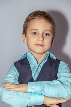 Young child with a confident expression wears a light blue shirt and dark vest, arms crossed. The child has short, light brown hair, and stands in a neutral pose against a plain backdropの写真素材