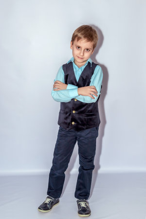 Young boy with light brown hair, wearing a blue shirt, navy vest, and pants, crosses his arms with a confident expression, standing with a slight tilt, in casual shoesの写真素材