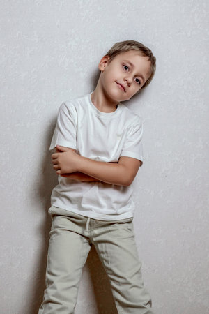 Young boy with short hair stands against a light textured wall, wearing a plain white t-shirt and light-colored pants, arms crossed, looking to the side. Neutral facial expressionの写真素材
