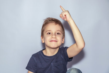Young child with short hair looks upwards, smiling with a raised index finger. Outfit includes a dark short-sleeve shirt. Light backdrop creates a soft contrast with the child's expressionの写真素材
