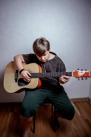 Young person sitting on a chair playing an acoustic guitar with focus and concentration, wearing...