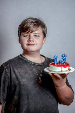 Young person holding small cake adorned with blue candles shaped as numbers 1 and 3, symbolizing age thirteen. Wearing dark T-shirt and pendant, expression is neutral and confidentの写真素材