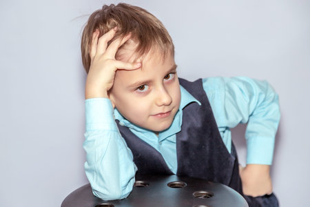 A young boy with light brown hair rests his head on his hand, wearing a blue shirt and dark vest,...