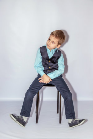 Young boy in a light blue shirt and dark vest seated on a stool, legs apart, looking to the side. He wears navy trousers and casual shoes, with a thoughtful expressionの写真素材