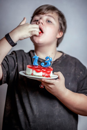 Boy enjoying cake, holding a plate with cake topped by blue candles shaped like numbers 1 and 3,...