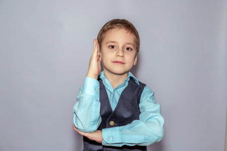 Young boy in a light blue shirt and dark vest poses with one hand touching his face, exuding confidence and charm. He has a focused expression and stands against a plain backgroundの写真素材