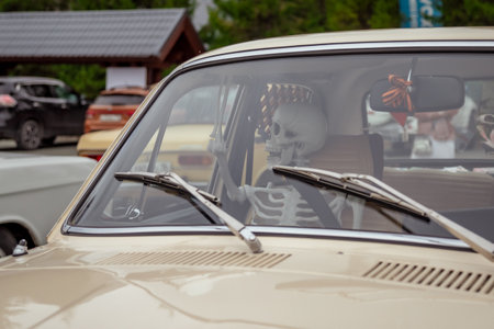 Skeleton seated in an old beige car parked outdoors with other vintage vehicles around. The skeleton appears to be driving, visible through the windshield with wipersの写真素材