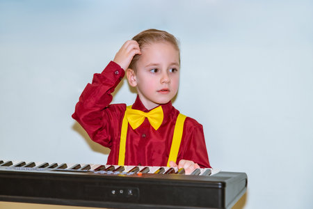 Young child in a red shirt with yellow bow tie and suspenders, standing by a keyboard, looking thoughtful. The child touches their head in a pondering gestureの写真素材