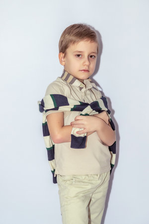 Young boy with short light brown hair wearing a beige shirt with a dark-stripped sweater draped around shoulders, holds a piece of fabric, looking at camera, pale light backgroundの写真素材