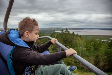 Child sitting on metal seat, looking at distant lake and forest under cloudy sky. He wears a backpack and casual attire, seemingly deep in thought while observing the expansive viewの写真素材