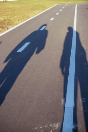 Shadows of two people stretch across a paved road with white lane markings under a bright sky. The perspective suggests a sunny day, with the figures elongated on the surfaceの写真素材
