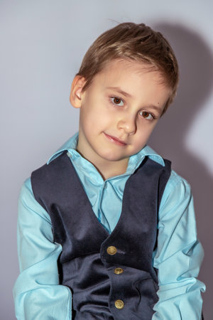 Young boy in light blue shirt and dark vest looks slightly sideways with a soft expression. Shadow falls to one side. Boy's hair is short and neatly combinedの写真素材
