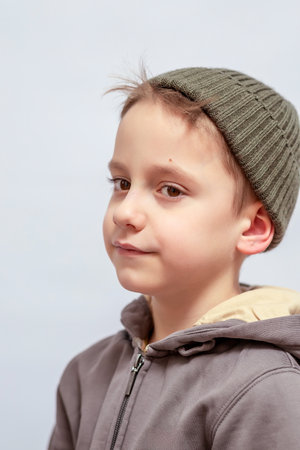 Young child with a thoughtful expression wears a gray hoodie and green knitted beanie, standing in profile. The close-up captures their curious gaze and youthful innocenceの写真素材