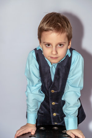 Young boy with short hair wears a light blue shirt under a dark vest with buttons, leaning forward on a surface, casting a shadow on a neutral backdropの写真素材
