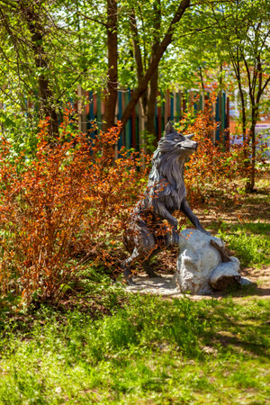 Wolf sculpture depicted in a garden, surrounded by sunlit trees and vibrant foliage. The scene captures a serene, natural setting. Shadows accentuate the detailed features of the statueの写真素材
