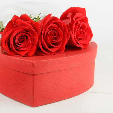 Gift box in the form of heart and scarlet roses on white wooden background. In the backgrounds a bow from organza.の写真素材