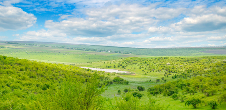 River valley with hills and terraces. On the slopes grow shrubs, trees and grass. In the blue sky, light cumulus clouds. Wide photo.の写真素材
