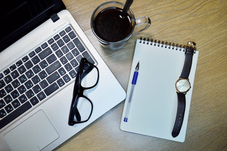 Mix of office supplies and gadgets on a wooden table background. View from above. Wrist Watch, laptop, pen, notebook, coffee and glasses on wooden backgroundの写真素材
