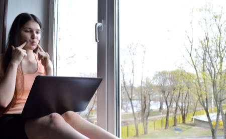 Young beautiful girl with dark hair with a laptop on the windowsillの写真素材