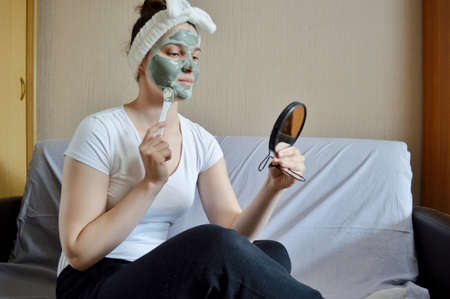 A young beautiful girl with dark hair with a white bandage on her head sits with a mirror in her hand and makes a glazed mask on her face. Spa at home.の写真素材
