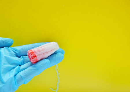 female hand in blue rubber glove holding a used gynological tampon on a yellow backgroundの写真素材