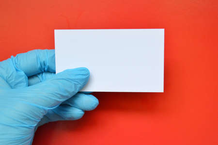 A hand in a blue rubber glove holds a white blank paper on a red background.の写真素材