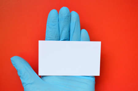 A hand in a blue rubber glove holds a white blank paper on a red background.の写真素材