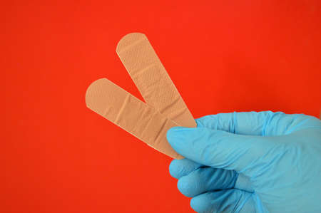 Female hand in blue rubber gloves holds two plasters on a red background.の写真素材