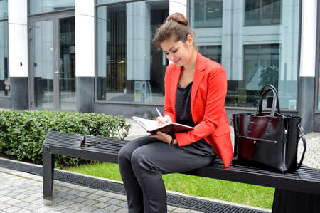 Beautiful business woman on the background of the modern office. A girl in a black overalls and a red jacket with a diary in her hands sits on a bench near the business centerの写真素材