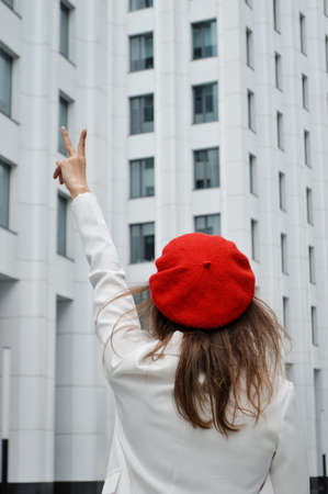 Beautiful business woman on the background of the modern office. The girl with dark hair in a white jacket and a red beret with his hand raised near the business center. Photo from the backの写真素材