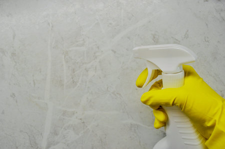 A hand in a rubber in a yellow glove holds a spray on a tile background