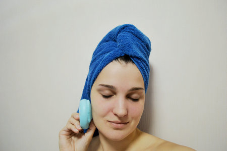 Woman with a blue towel on her head on a light background cleans her face with an electric brush