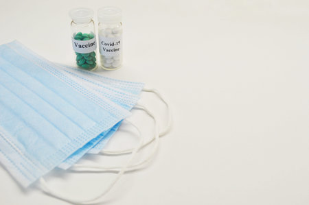 Glass jars with vaccine and medical masks on a light background.