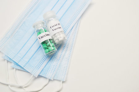 Glass jars with vaccine and medical masks on a light background.