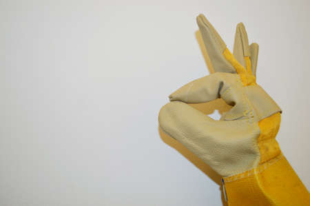 Hand in beige and yellow work cloth glove shows ok sign against white wall backgroundの写真素材
