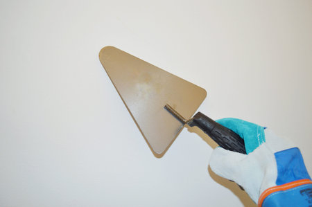 Hand in blue work cloth glove holds a metal trowel against white wall background