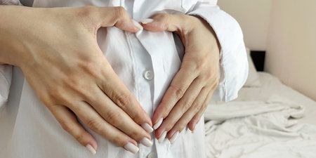 Pregnant girl in a white shirt holds her hands on her stomach in the shape of a heart.