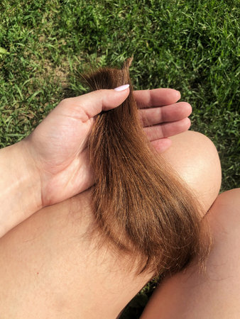 Hand holding a long strand of brown hair, resting on a leg, surrounded by green grass, showing natural beauty and hair texture in a serene outdoor settingの写真素材