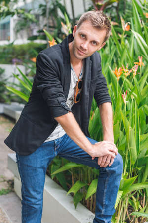 Young guy ina black jacket and jeans. Handsome smart man standing outside with green area on background. Vertical photo.の写真素材