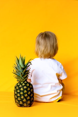 A 2-year-old child with blond hair in a white t-shirt and yellow pants with a ripe big pineapple on a yellow background. emotions The concept of childhood and family. Front view of child on yellow isolated background with happy and excited expression.の写真素材