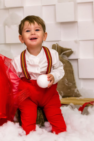 A little boy in red clothes have fun in the artificial snow by the fireplace in the studio. A little kid in red christmas costume having fun beside a decorated christmas tree. Baby playing with falling artificial snow flakes near a christmas tree. A kid playing with artificial snow flakes falling near a christmas tree. Christmas concept. High quality photoの写真素材
