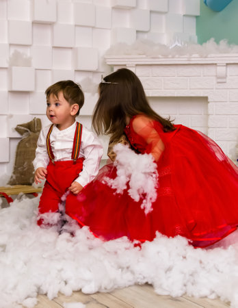 A little girl and a boy in red clothes have fun in the artificial snow by the fireplace in the studio. The girl is holding a box with gifts. Little kids in red christmas costume having fun beside a decorated christmas tree. Two kids playing with falling artificial snow flakes near a christmas tree.Kids playing with artificial snow flakes falling near a christmas tree. Christmas concept. High quality photoの写真素材