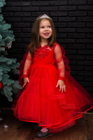 Little girl in red dress smiling by Christmas tree. Little beautiful girl in a red evening dress the Christmas tree. Girl in red in Christmas decorations. Christmas concept. New Year. Baby near a Christmas tree. High quality photoの写真素材