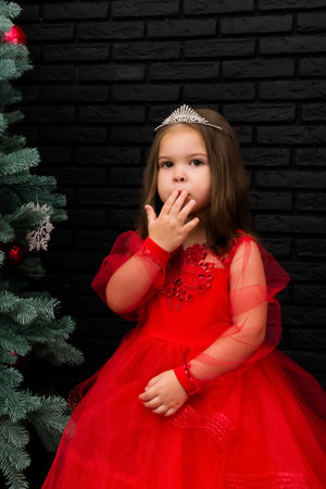 Little girl in red dress smiling by Christmas tree. Little beautiful girl in a red evening dress the Christmas tree. Girl in red in Christmas decorations. Christmas concept. New Year. Baby near a Christmas tree. High quality photoの写真素材